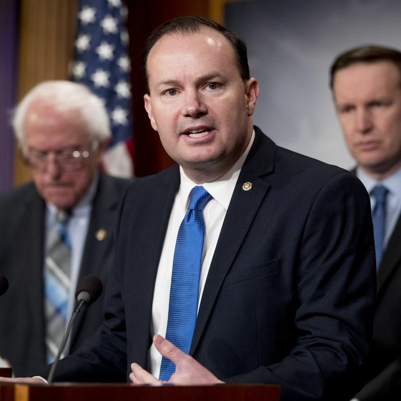Three men in suits stand at podiums. The man in front speaks, gesturing with one hand, while the two men behind him listen. An American flag is visible in the background.