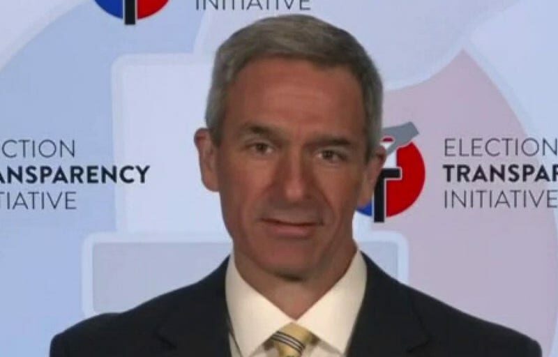 A man in a suit and tie stands in front of a backdrop that reads Election Transparency Initiative, with circular logos visible in the background.