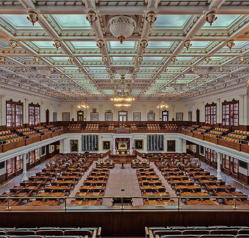 Large, ornate legislative chamber with rows of wooden desks and chairs, high coffered ceiling with chandeliers, large windows, and balconies with additional seating along the sides and back.