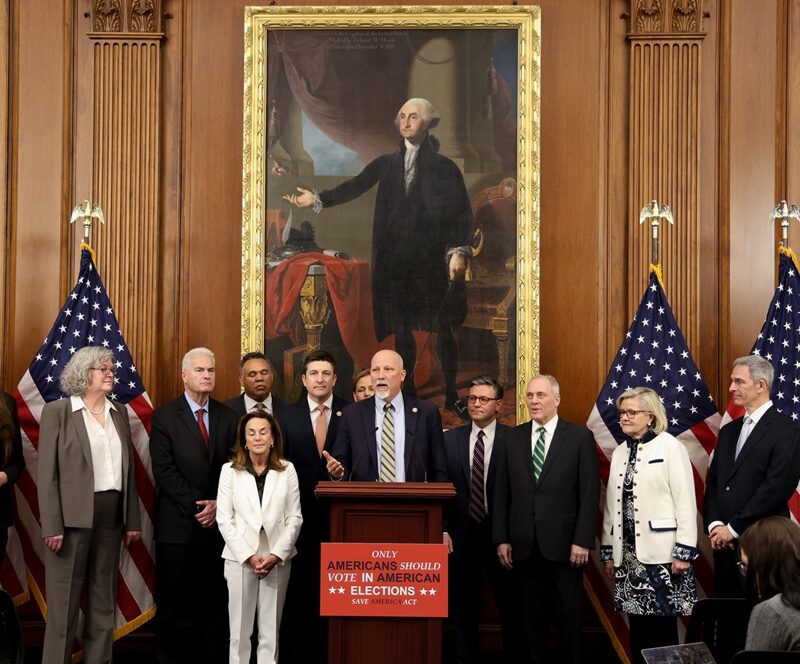 A group of people stand in a formal room with four U.S. flags, a large portrait of George Washington, and a sign reading Only Americans Should Vote in American Elections. Ken Cuccinelli speaks at the podium during the SAVE Act press release.