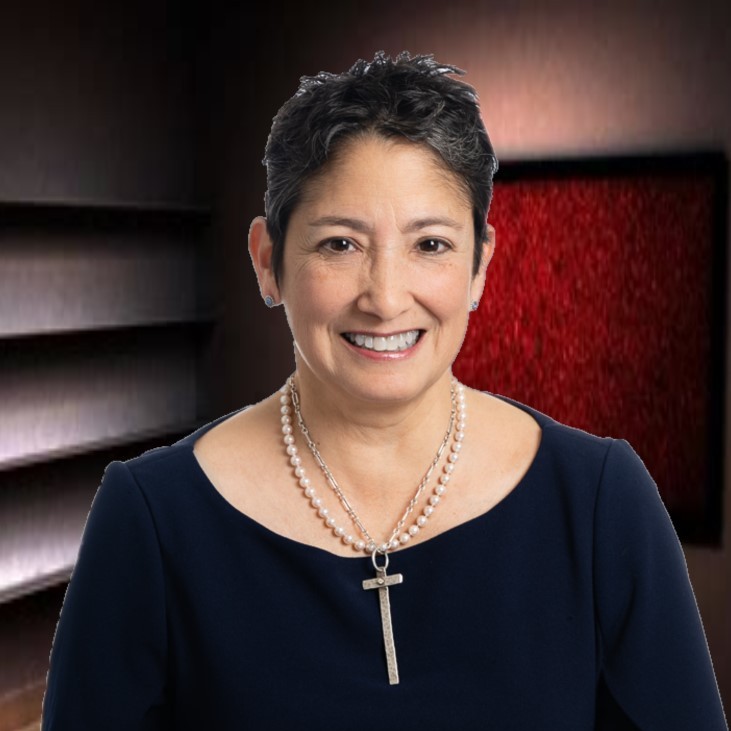 A smiling woman with short dark hair, wearing a navy blue top, a double-strand pearl necklace with a cross pendant, standing in a dimly lit room with shelves and red artwork on the wall.